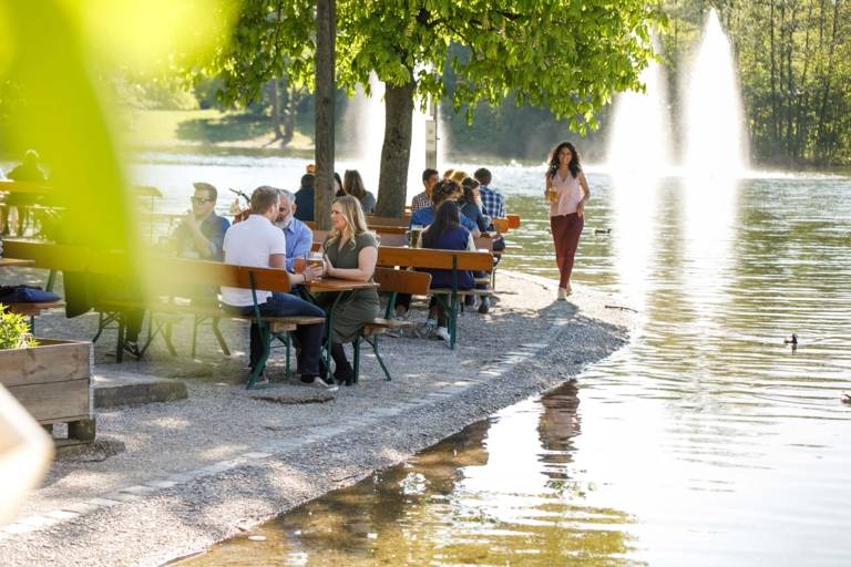 A lot of persons are sitting on benches in a beer garten in Munich at a lake and are talking to each other. In the foreground is a green leaf.