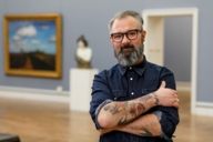 A man with beard, glasses and a tattooed forearm is standing in the Neue Pinakothek in Munich.