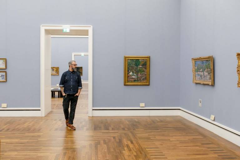 A man with glasses and beard is going through the Neue Pinakothek in Munich and is looking at the paintings.