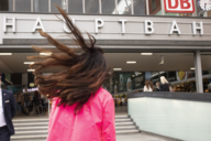 A woman with a pink mackintosh and flying hair walks towards the entrance of a main station.