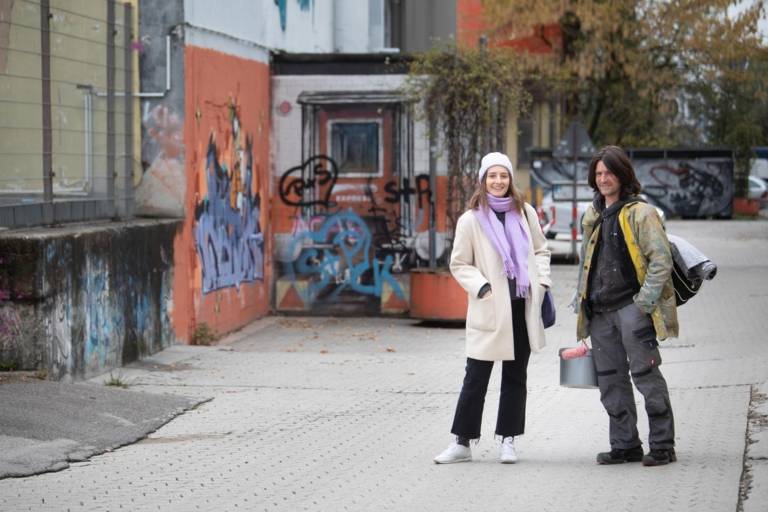 A woman in a coat and an artist with a paint bucket smile into the camera in Munich.